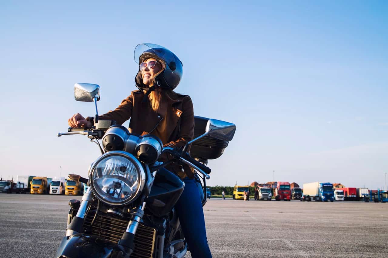 Mulher sorridente de capacete e óculos escuros sentada em uma motocicleta preta, com caminhões estacionados ao fundo sob céu azul.