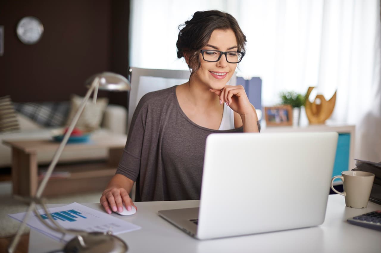 Mulher jovem de óculos sorrindo enquanto usa um notebook em uma mesa de escritório, em casa. Ela está pesquisando como simular financiamento de energia solar.