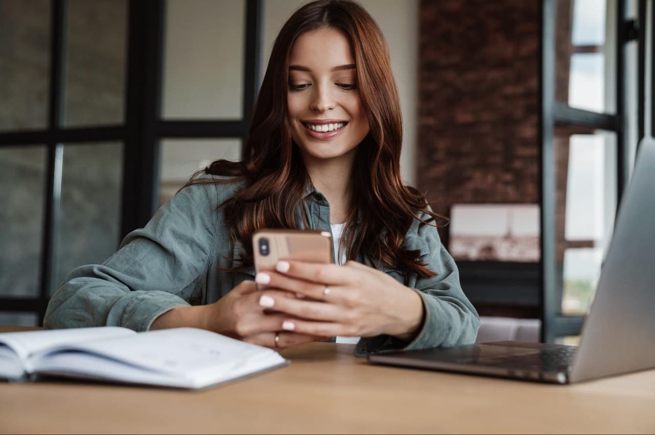 Mulher sorridente de cabelos lisos castanhos, vestindo jaqueta jeans e acessando saque-aniversário do FGTS pelo celular.