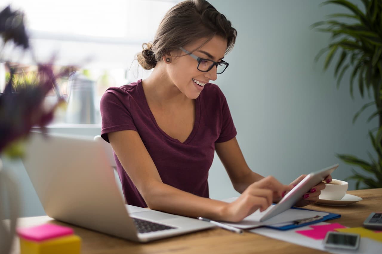 Mulher jovem de óculos sorrindo, enquanto usa um tablet para pesquisar sobre como tirar os planos do papel. Ela está em uma mesa de trabalho, onde também há um laptop, folhas de papel, caneta e uma xícara de café.