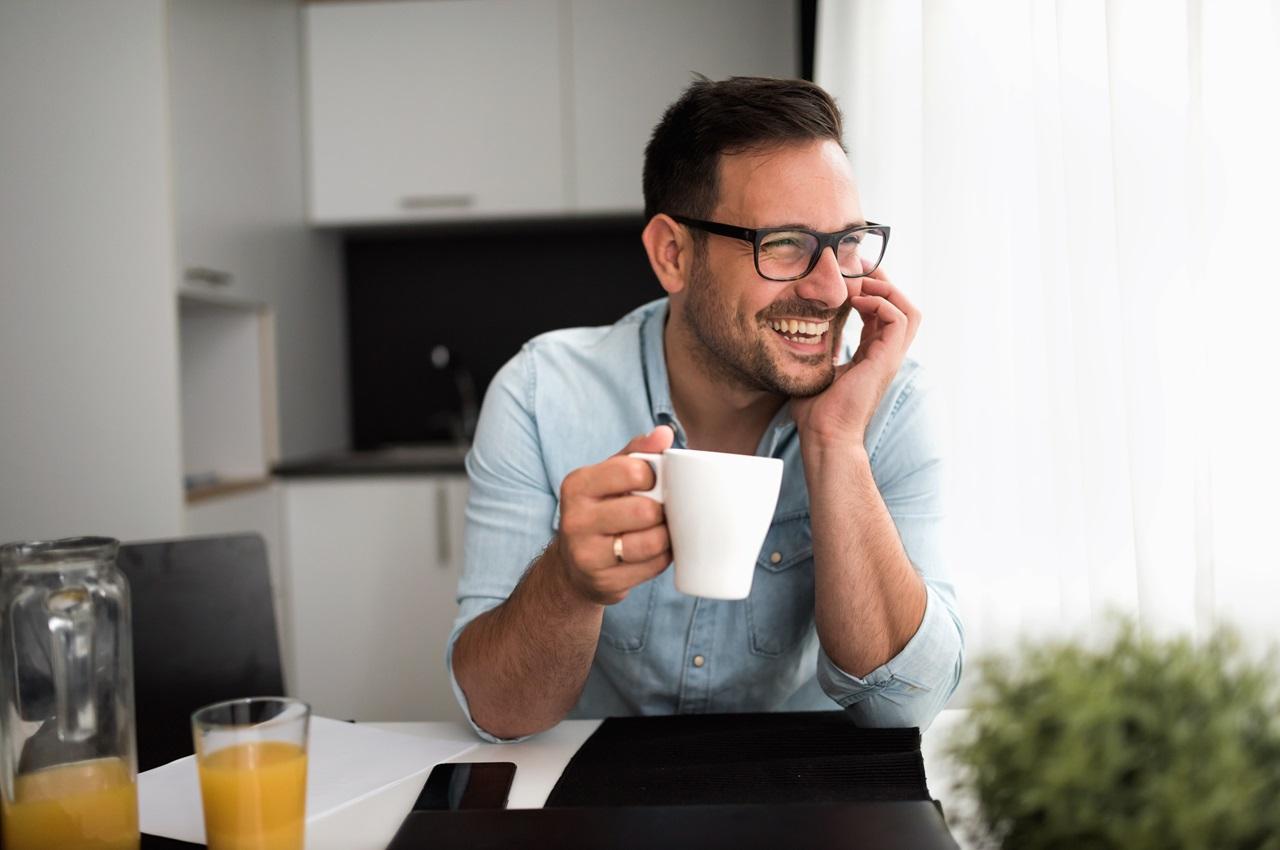Homem sorridente segurando xícara de café e olhando pela janela da sua cozinha.