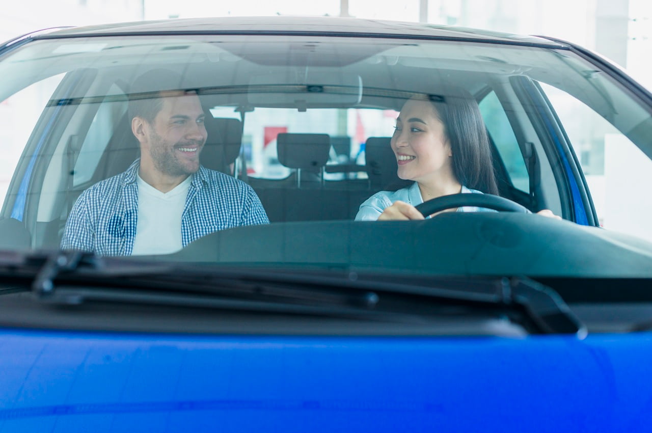 Pessoas sorrindo dentro de um carro azul. Uma delas está na direção do veículo.