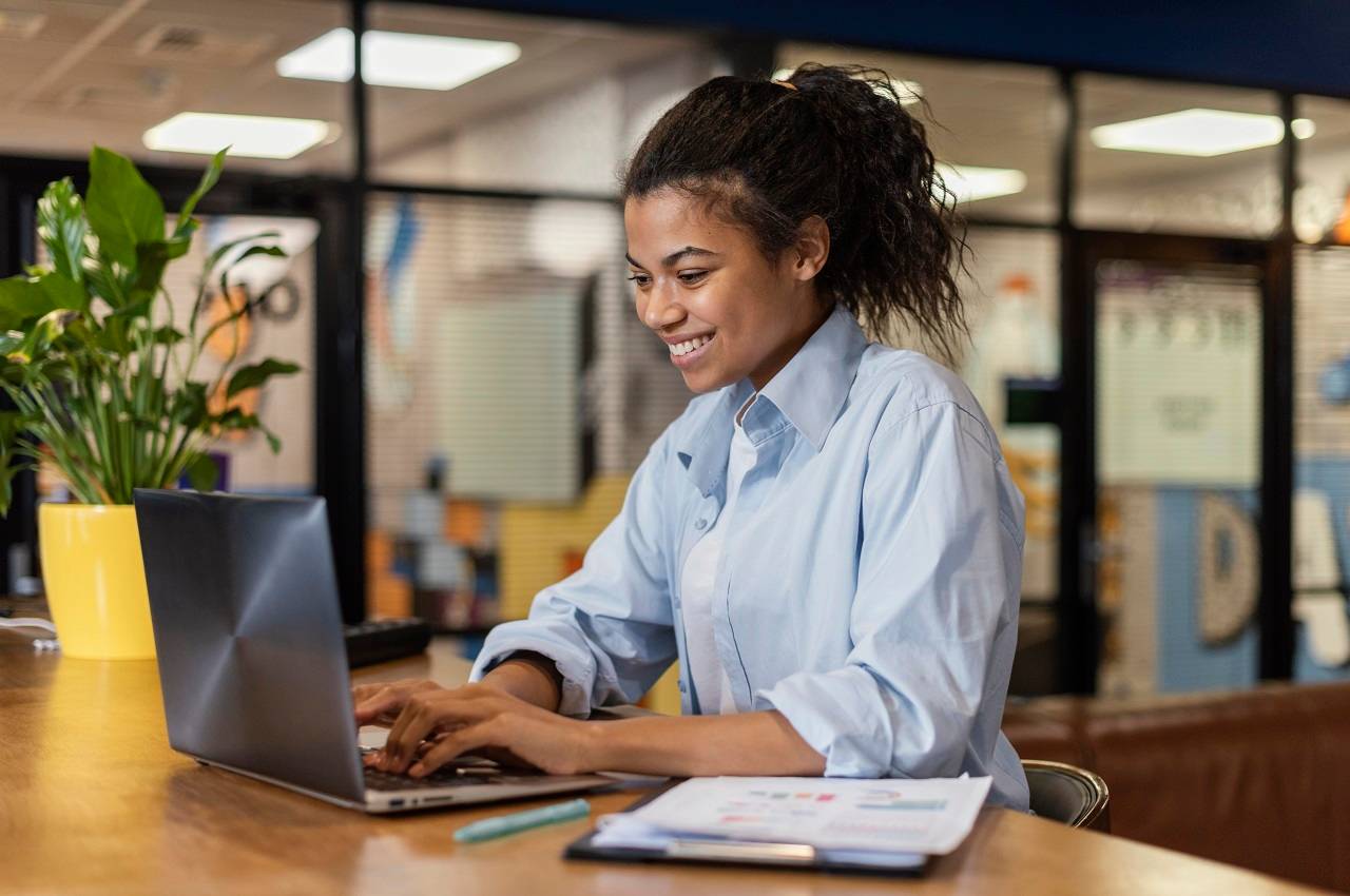 Mulher sorridente vestindo blusa social azul clara e ditando no laptop do seu trabalho. Ao seu lado tem uma planilha com gráficos.