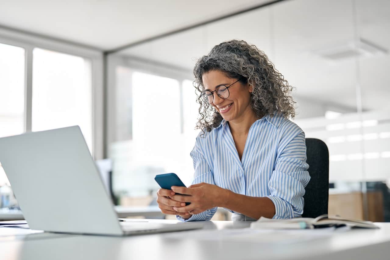 Mulher sentada, com uma mesa em frente, com notebook e segurando um celular nas mãos