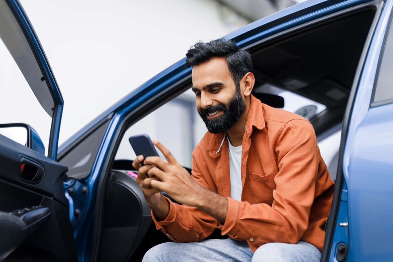 Homem sorrindo, sentado dentro do carro com a porta aberta, segurando um celular