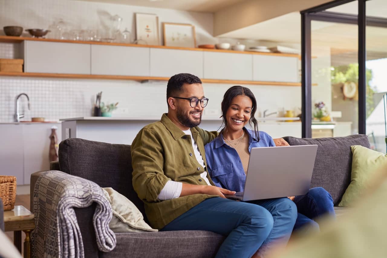Casal sorrindo sentado no sofá com notebook apoiado nas pernas.
