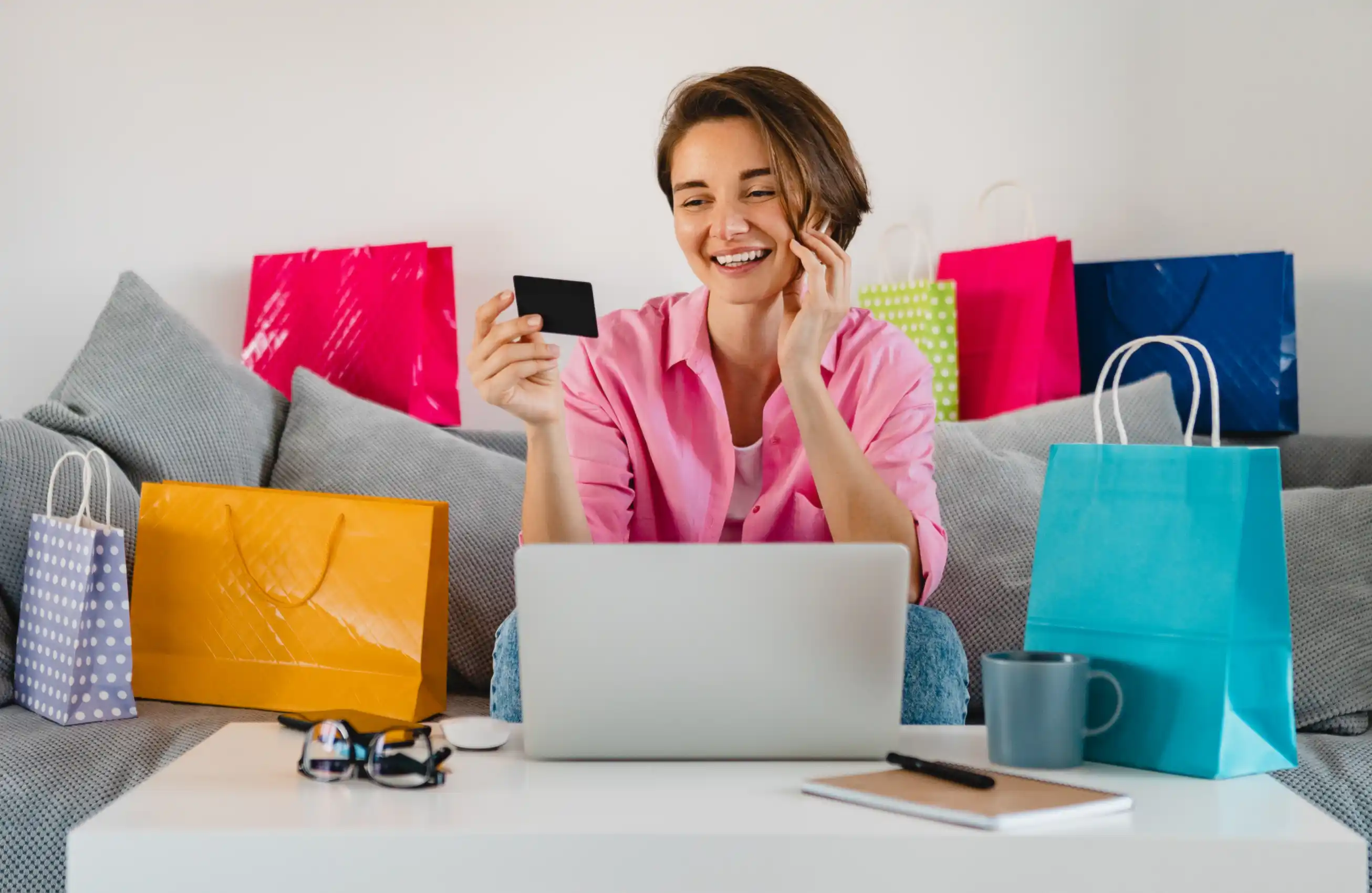 Mulher sorridente sentada no sofá com várias sacolas de compras ao redor, segurando um cartão de crédito na mão direita e usando um laptop apoiado no colo.&nbsp;