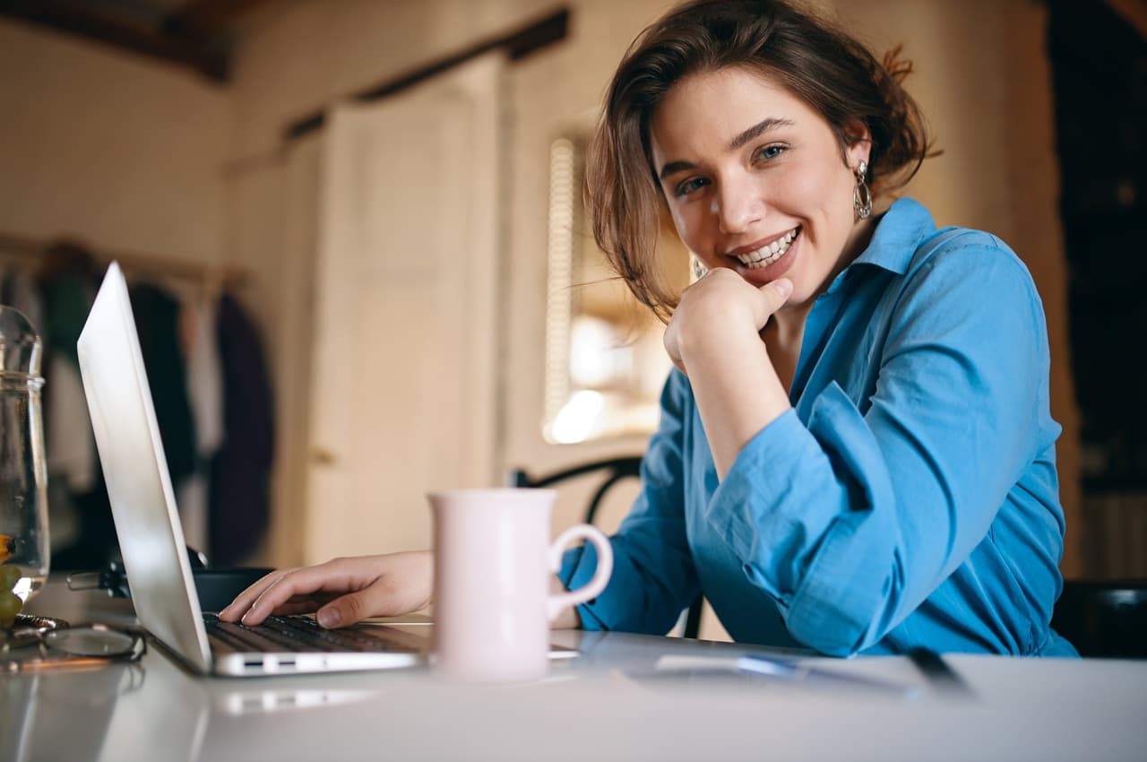 Mulher sorridente de camisa azul apoiada na mão enquanto usa um notebook em uma mesa.