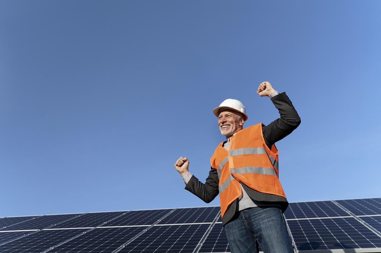 Homem sorridente, usando capacete e colete de segurança, em frente a painéis solares sob um céu azul.