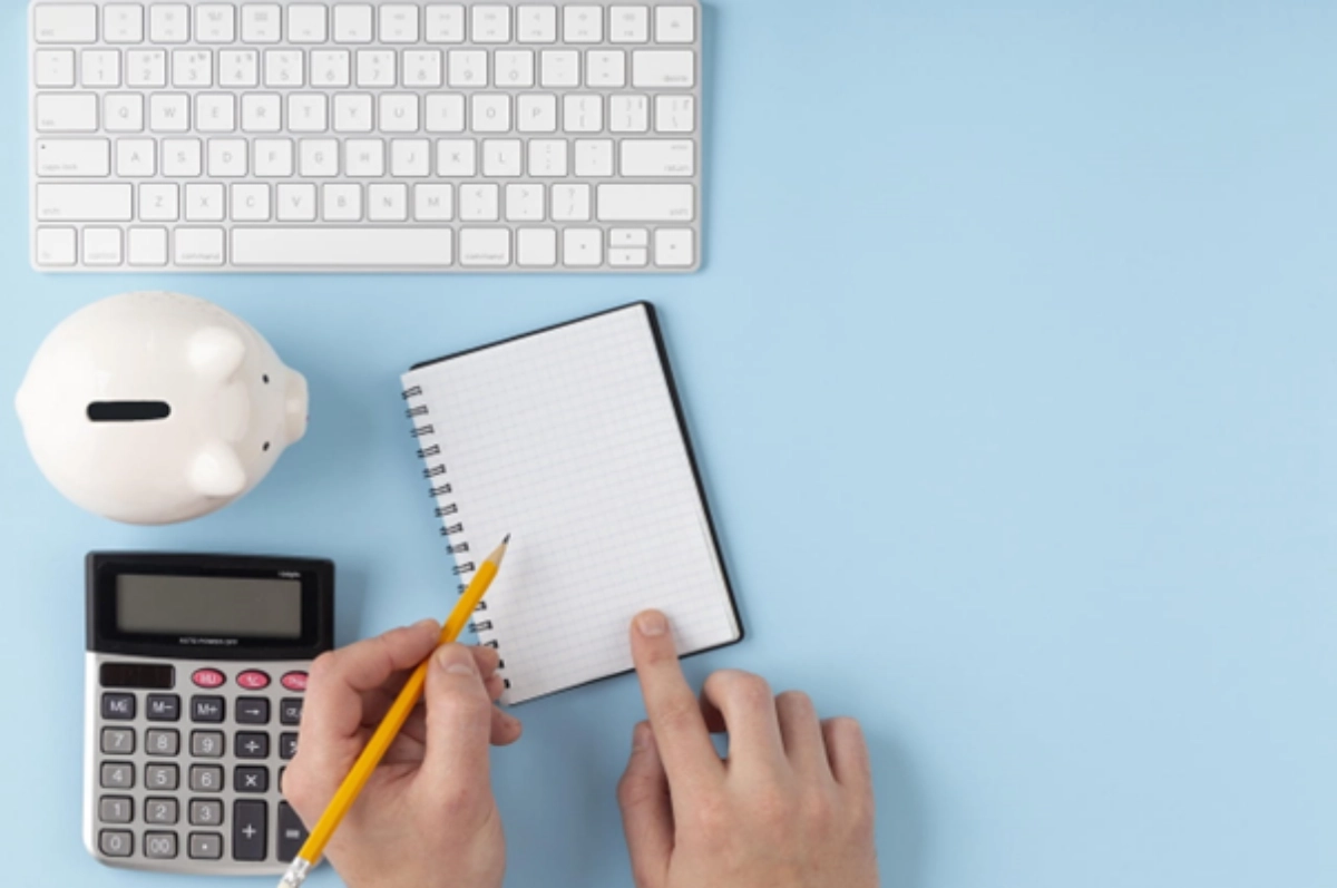 Mão segurando lápis amarelo, com calculadora, caderno de notas, cofre de porquinho e teclado sobre um fundo azul.