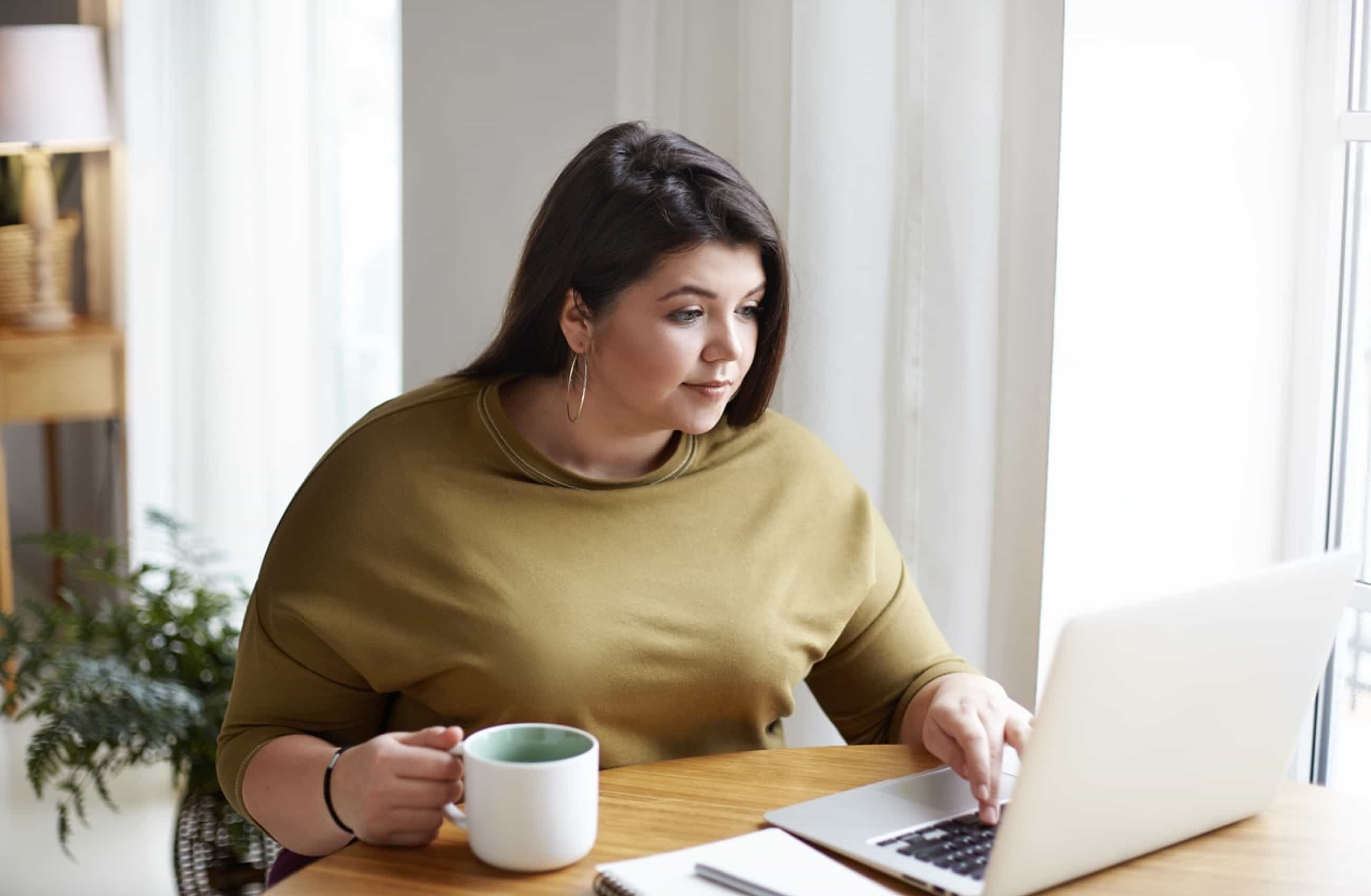 Mulher branca com camiseta bege está sentada em frente a mesa mexendo em um notebook enquanto segura uma caneca branca na mão.