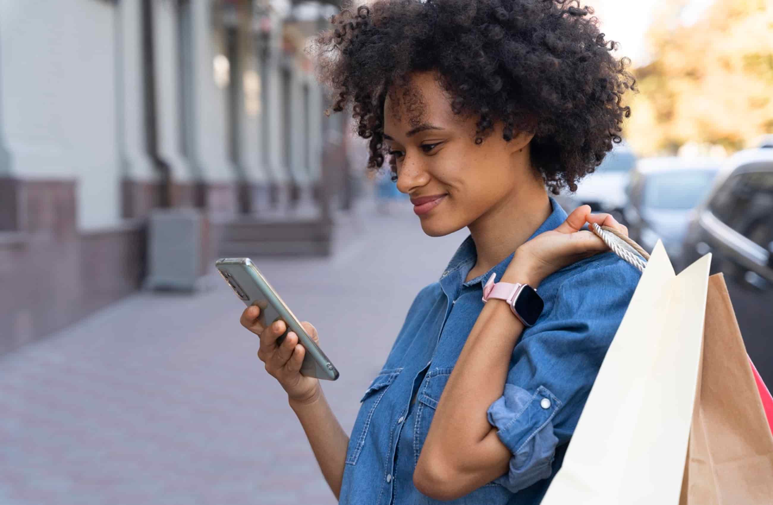 Mulher negra com uma camisa social jeans está segurando sacola de compras com uma das mãos, e na outra digita no celular. Ela está numa calçada da rua.