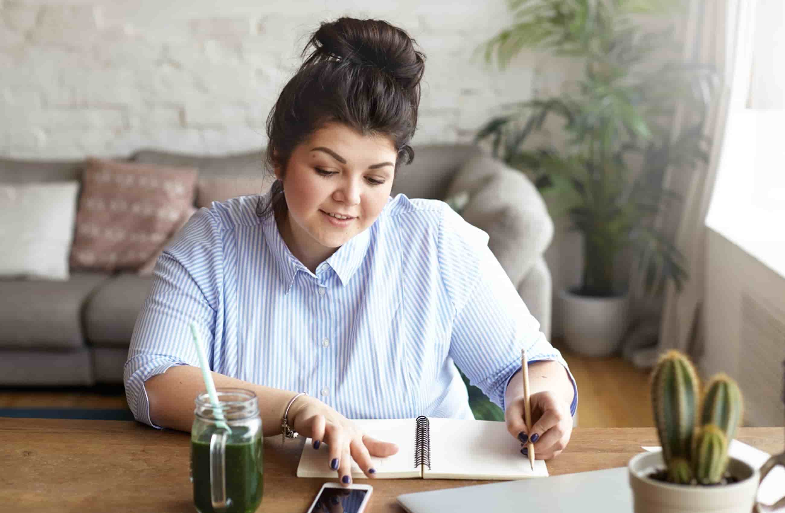 Mulher branca de camisa social listrada azul com um coque no cabelo, está sentada em frente a mesa escrevendo em um caderno.