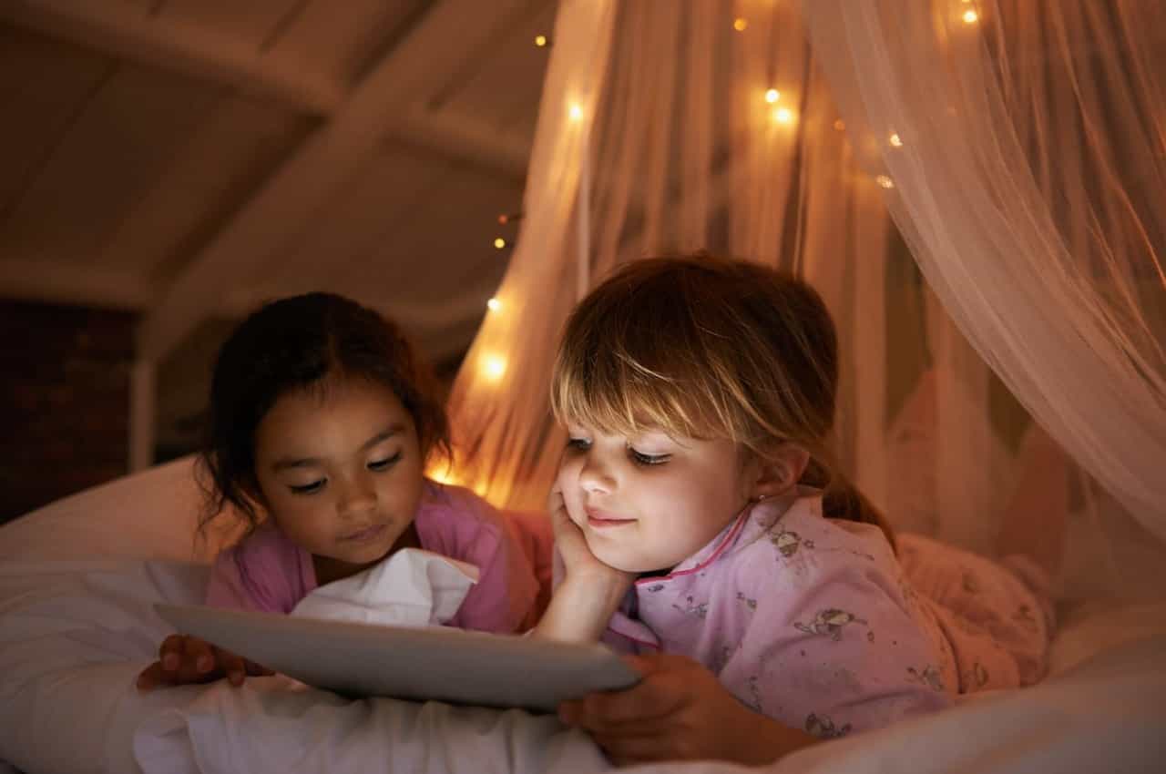 Duas crianças pequenas, vestindo pijamas de bichinhos e deitadas de barriga para baixo em uma cabana de lençóis lendo um livro.