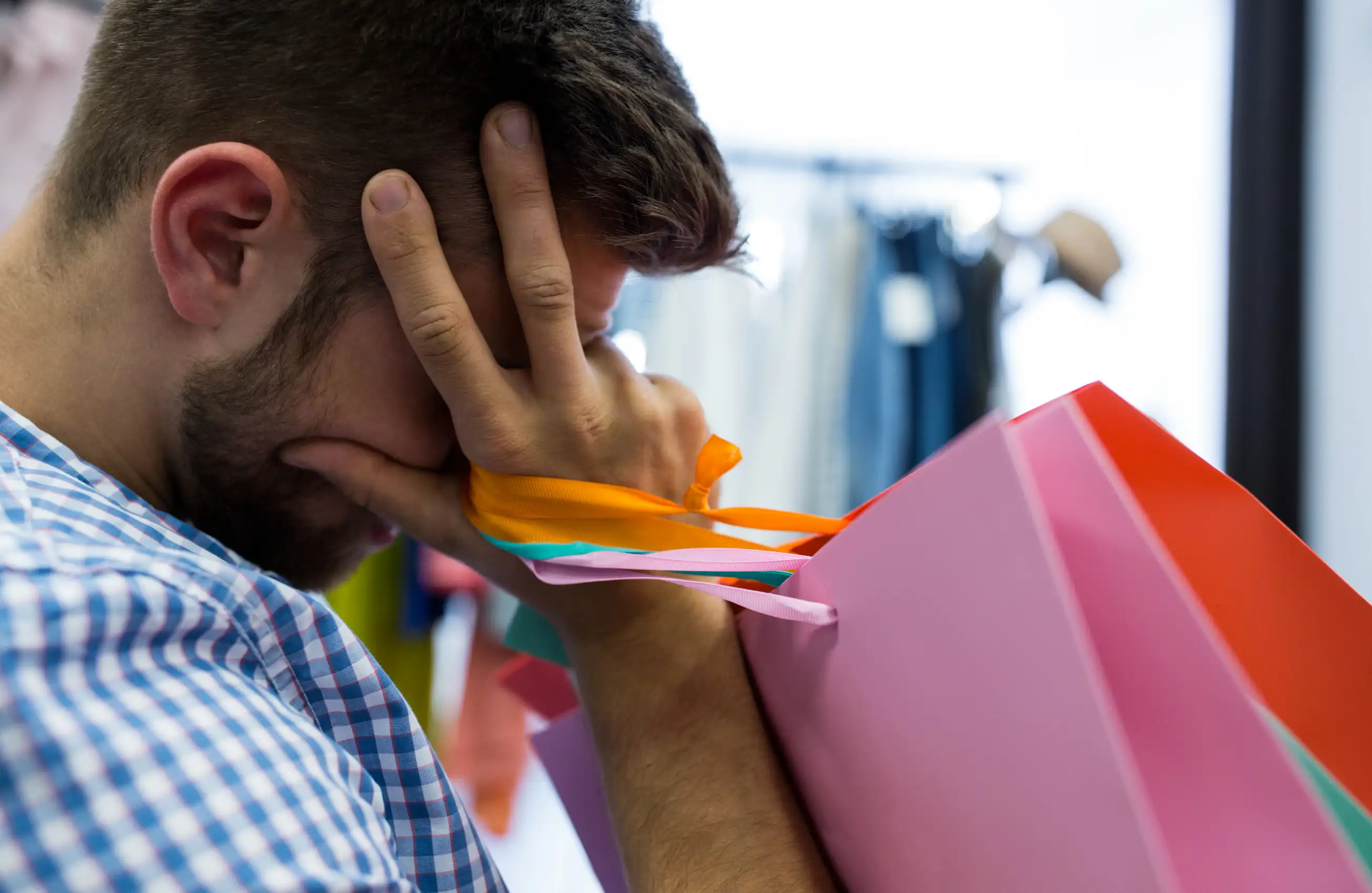 Homem segurando várias sacolas de papel coloridas sobre o ombro, com a cabeça entre as mãos em tom de frustração, ambiente de loja ao fundo.