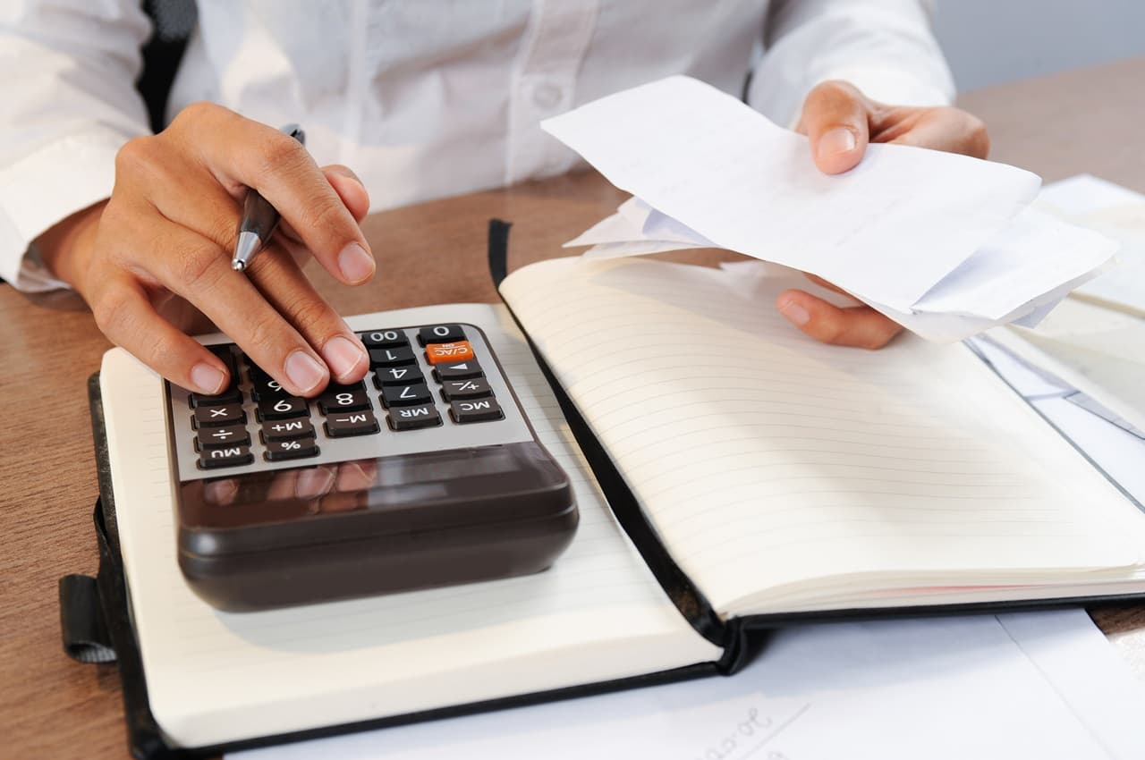Mãos usando calculadora e caderneta com notas fiscais para calcular despesas sazonais.