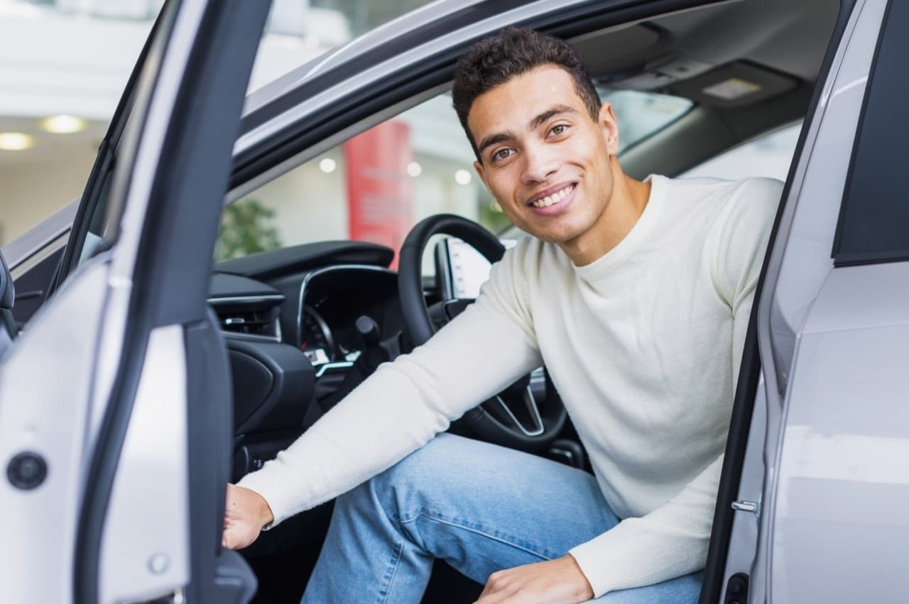 Homem jovem sorridente sentado no banco do motorista de um carro prata com a porta aberta em uma concessionária.