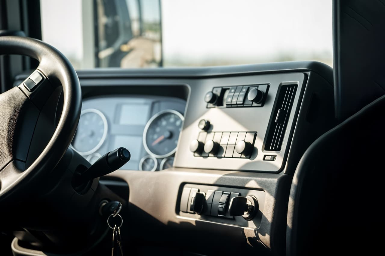 Interior da cabine de um caminhão, com foco no painel de instrumentos, volante e controles sob luz solar.
