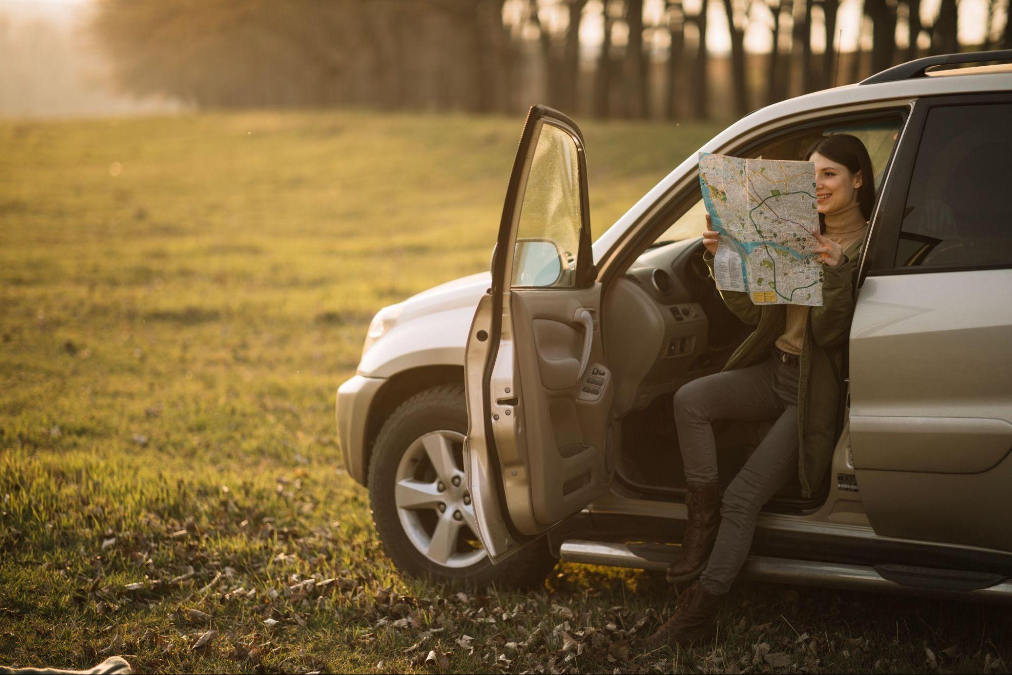 Uma mulher sentada em uma SUV prateada, com a porta aberta, segurando um mapa e sorrindo. O cenário é um campo verde sob a luz suave do sol, com árvores ao fundo.