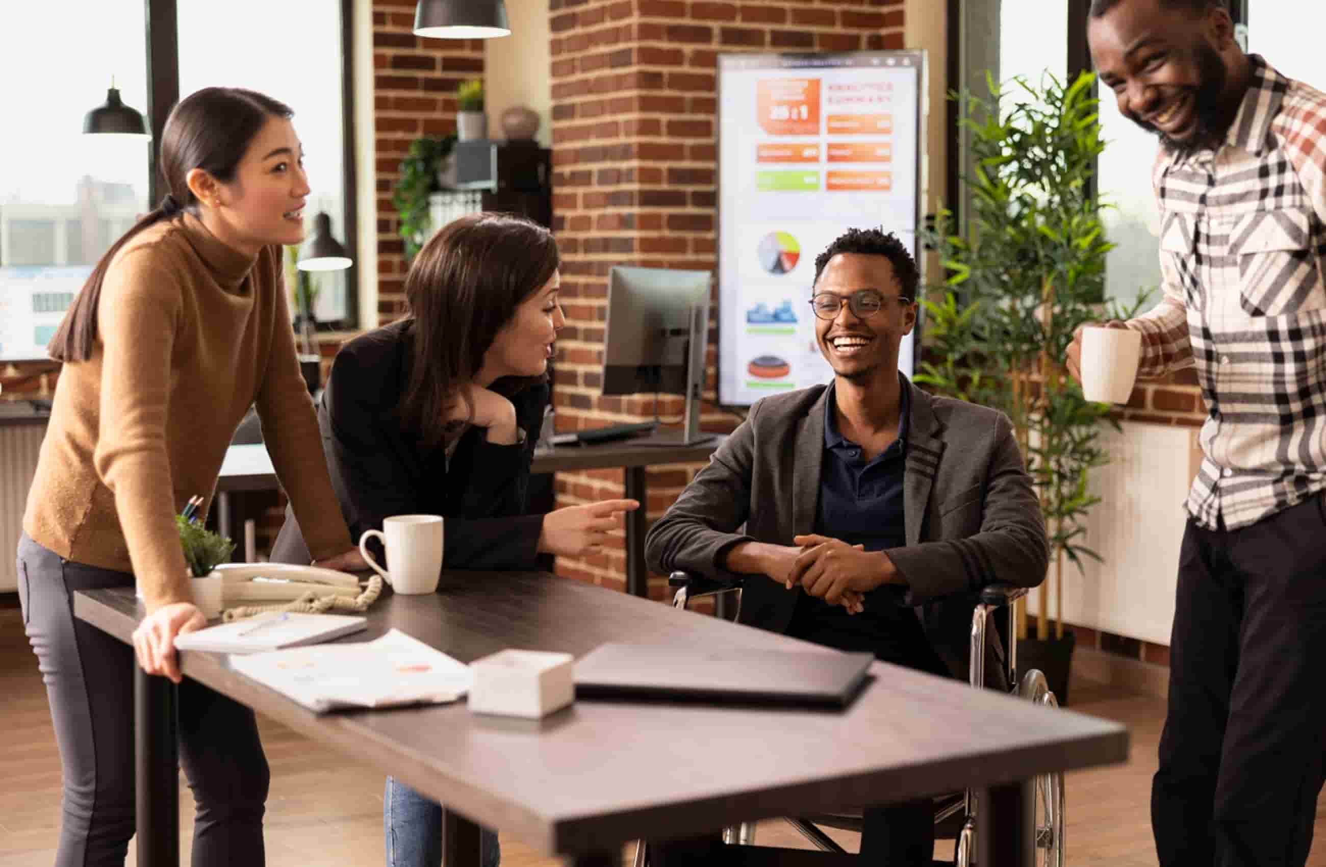 Quatro profissionais conversavam sorridentes em um escritório moderno. Uma mulher asiática está apoiada em uma mesa, dialogando com dois homens negros, sendo um deles cadeirante, e uma mulher branca que seguram canecas de café.