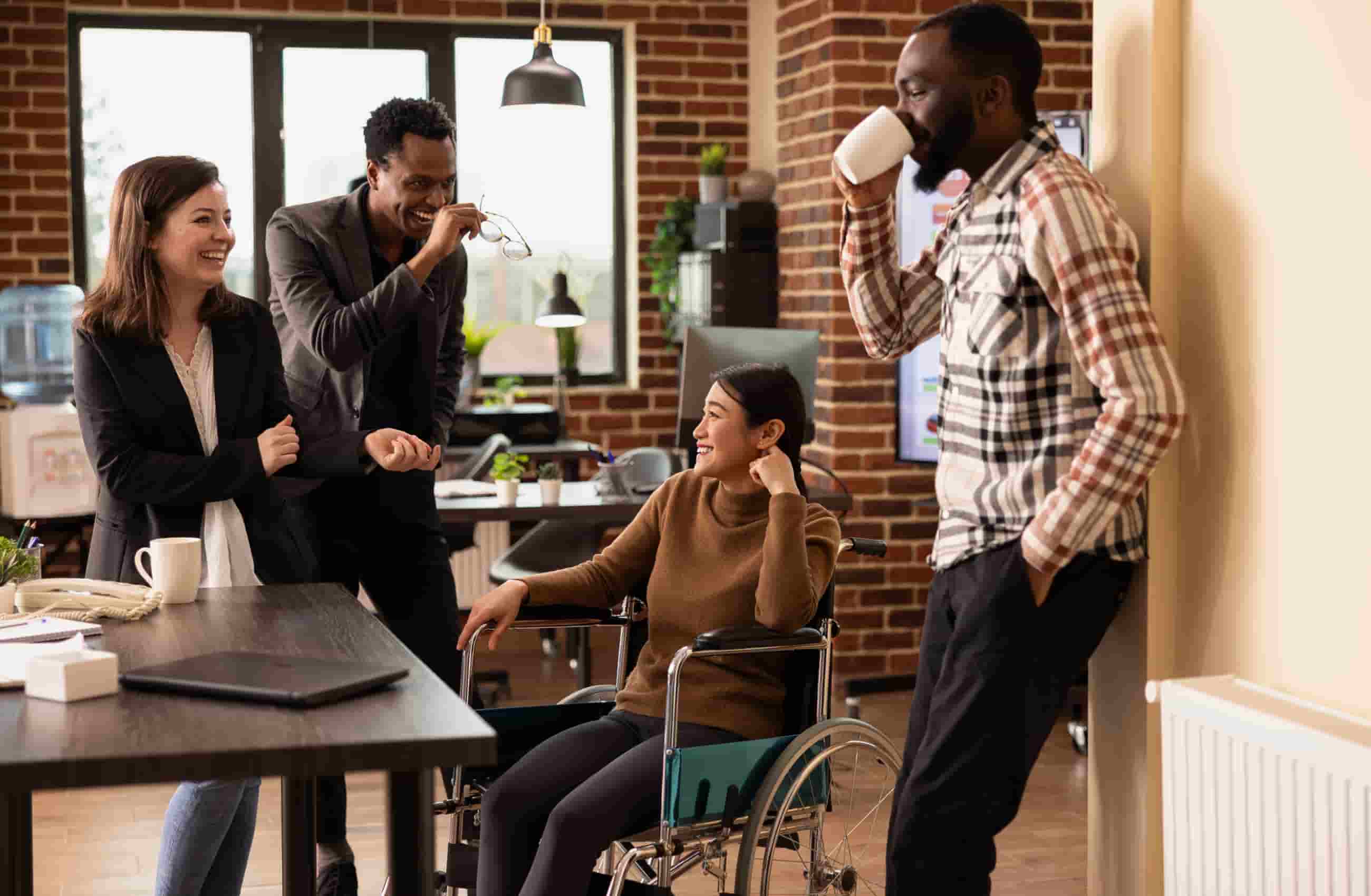 Quatro profissionais conversavam sorridentes em um escritório moderno. Uma mulher asiática cadeirante está no centro, dialogando com dois homens negros e uma mulher branca que seguram canecas de café. 