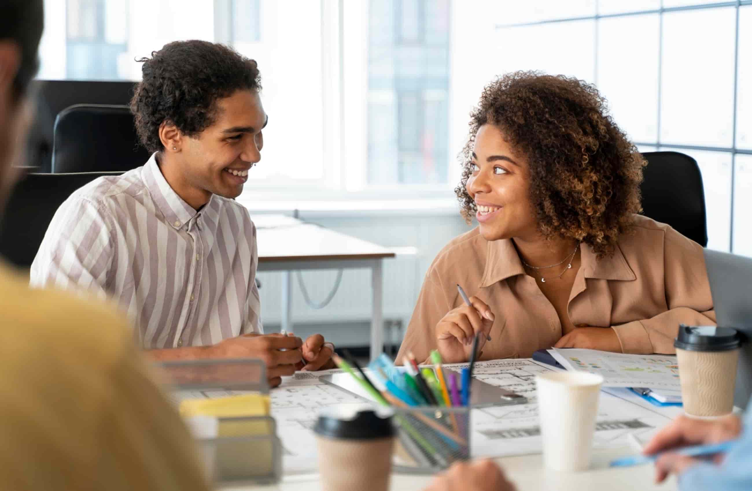 Um homem e uma mulher, ambos negros, estão vestidos com roupas sociais em um ambiente de trabalho. Estão sentados envolta de uma mesa com vários papéis. Ambos sorriem um para o outro. A imagem representa a diversidade nas empresas.