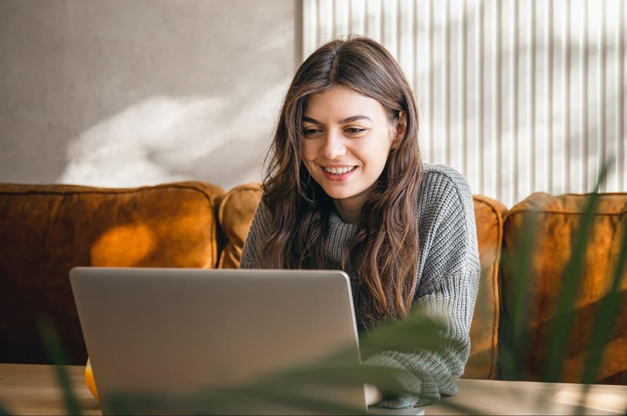 Mulher sorridente sentada em um sofá e utilizando um notebook. Ela está pesquisando sobre as vantagens do AOC.