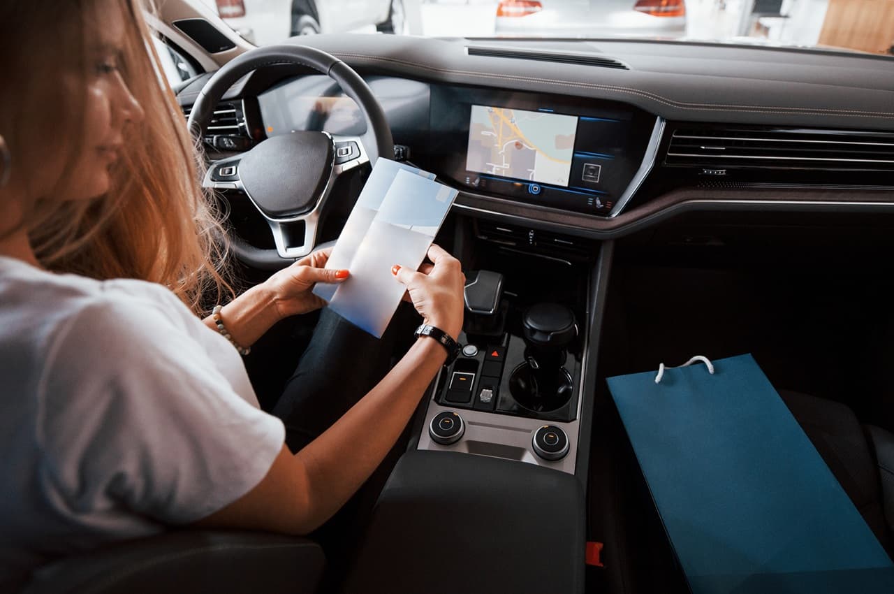 Mulher sentada no banco do motorista de um carro, examinando alguns documentos, com o painel digital visível ao fundo.