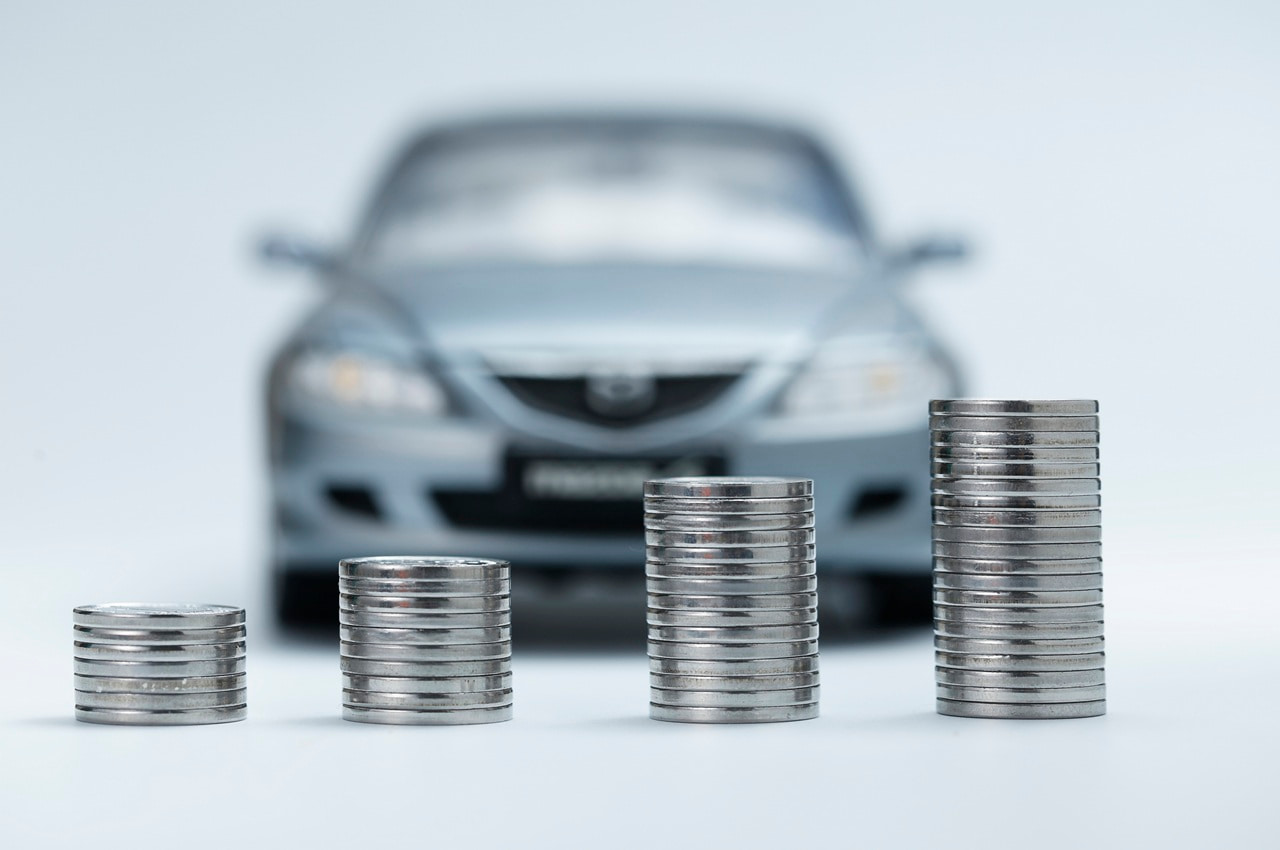Pilha de moedas crescendo na frente de um carro cinza desfocado ao fundo, simbolizando o empréstimo com garantia de veículo financiado.