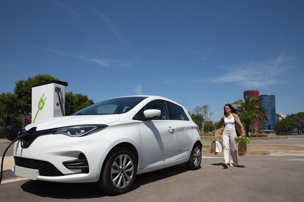 Mulher caminhando com sacolas de compras em direção ao seu carro elétrico branco, que está conectado a uma estação de recarga.