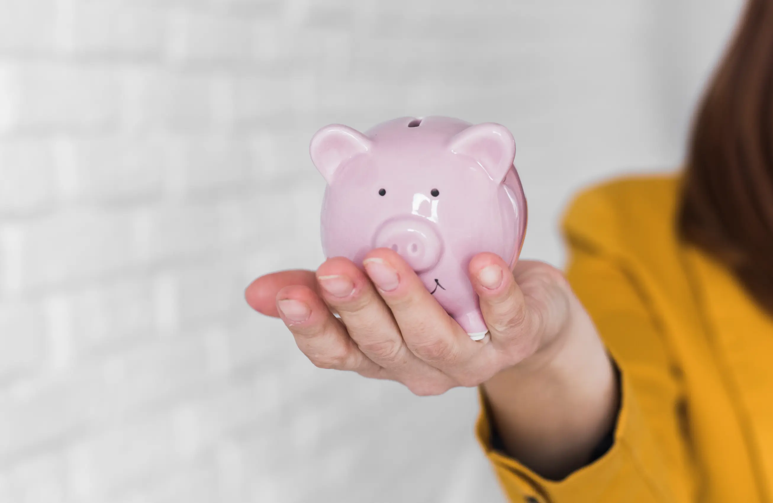 Mão estendida segurando um cofrinho em formato de porquinho rosa, com pessoa vestindo casaco mostarda parcialmente visível ao lado direito; fundo claro desfocado.&nbsp;