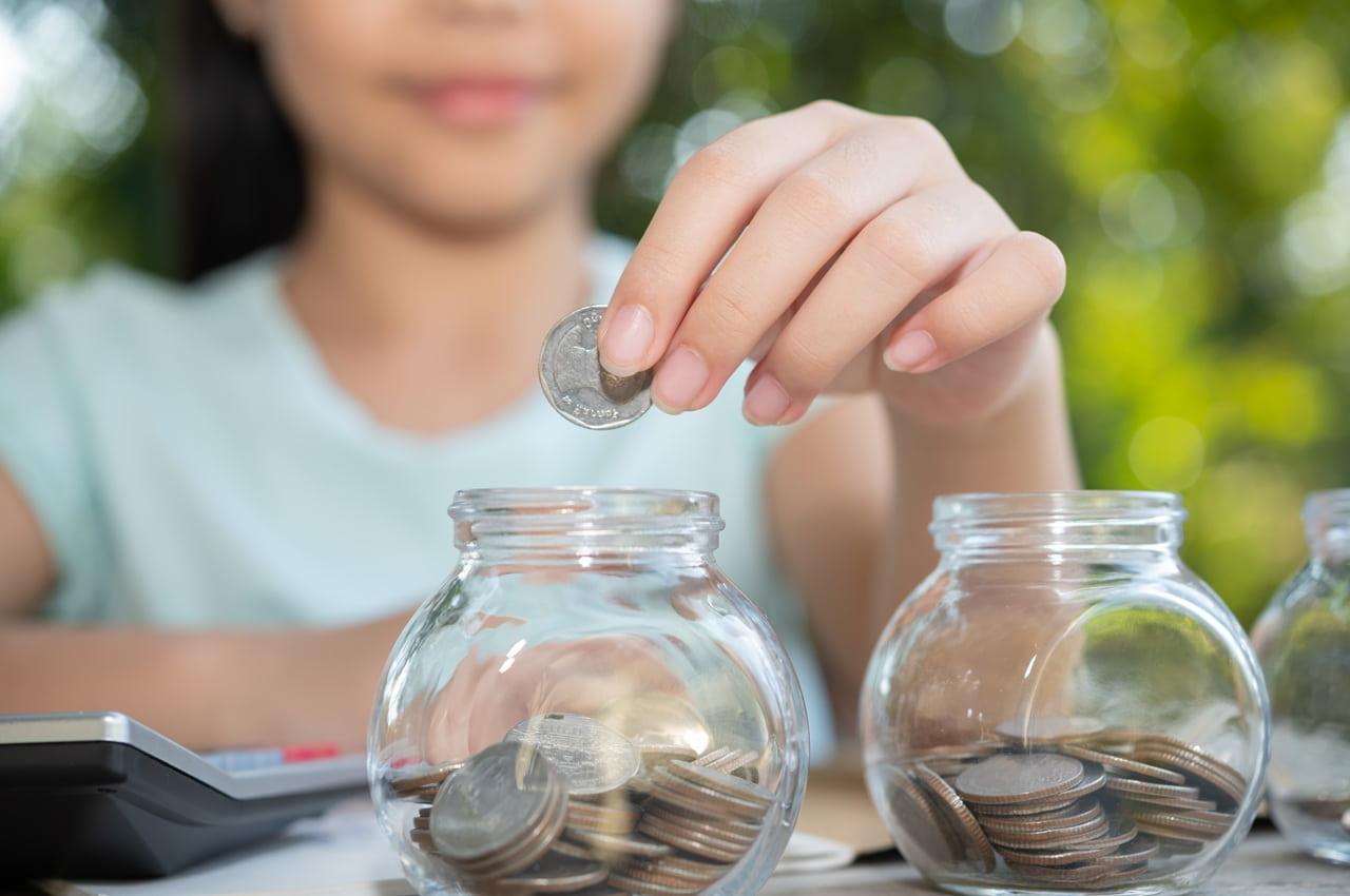 Criança colocando moedas em cofrinhos de vidro transparentes. Ela está aprendendo sobre educação financeira infantil.