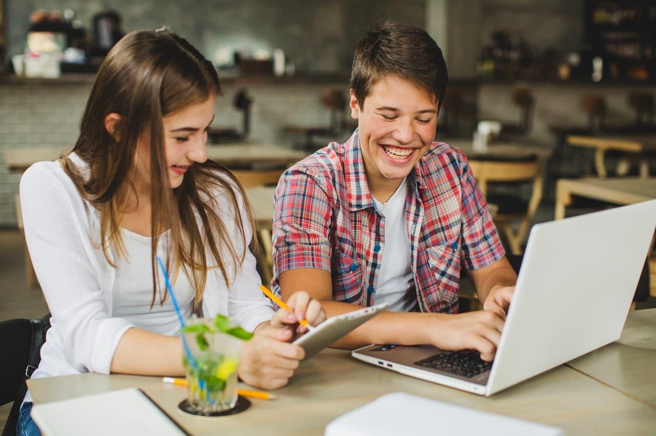 Dois adolescentes sorrindo enquanto usam um notebook e um tablet. Eles estão estudando sobre educação financeira para crianças e jovens.