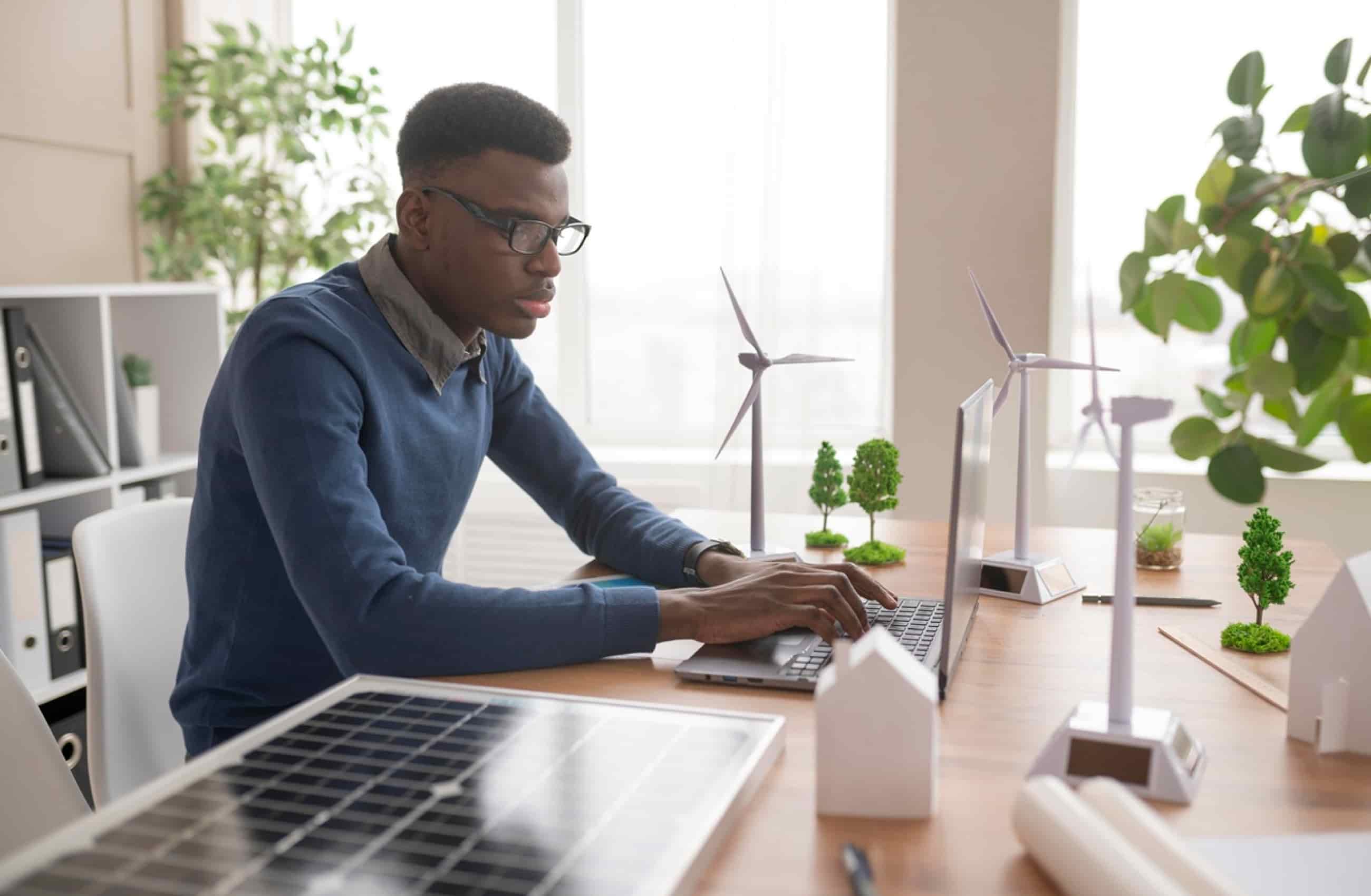 Homem negro usando camisa azul está sentado em uma cadeira enquanto trabalha em seu notebook sobre a mesa.