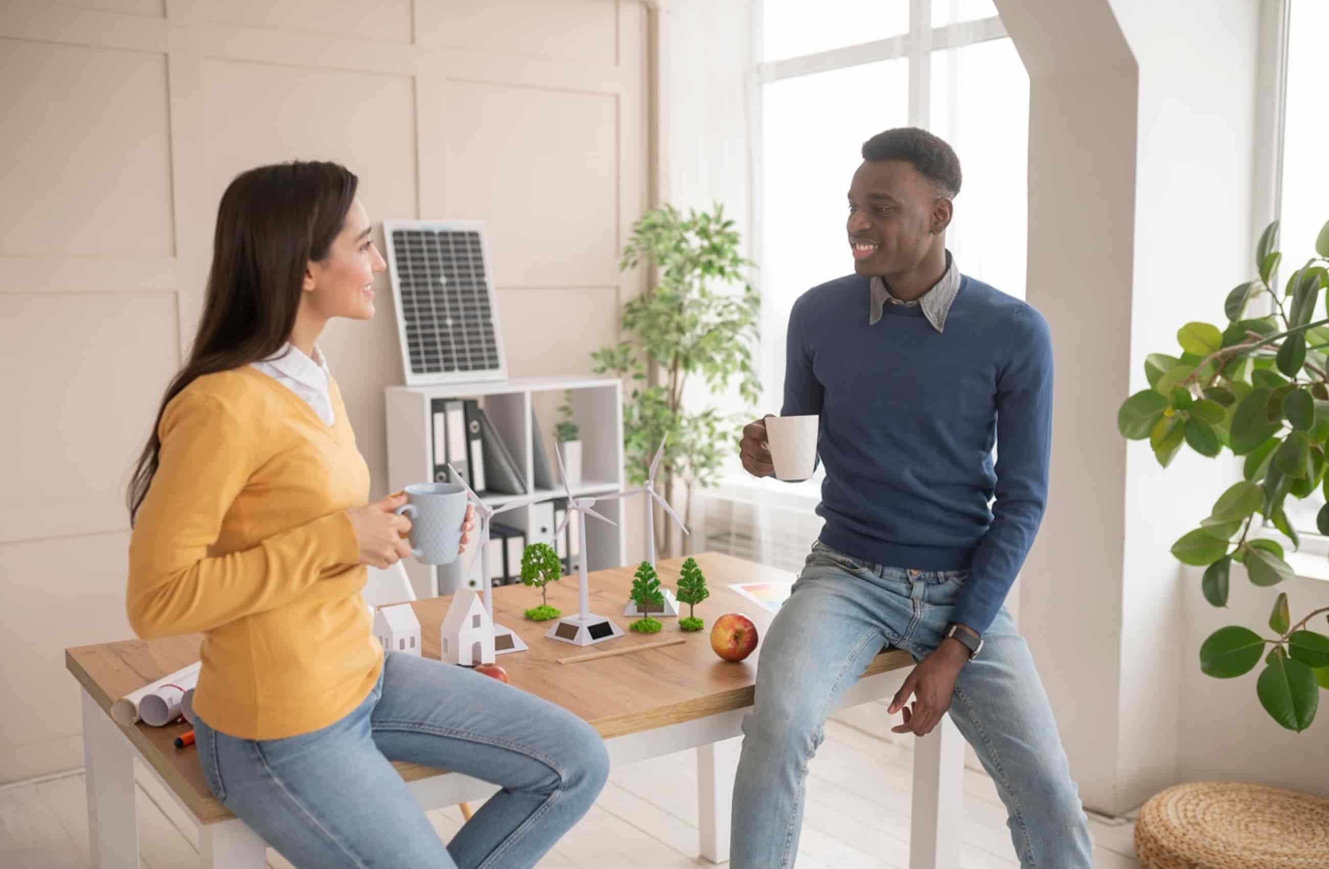 Homem negro e mulher branca estão conversando sentados em uma mesa, enquanto seguram uma caneca.