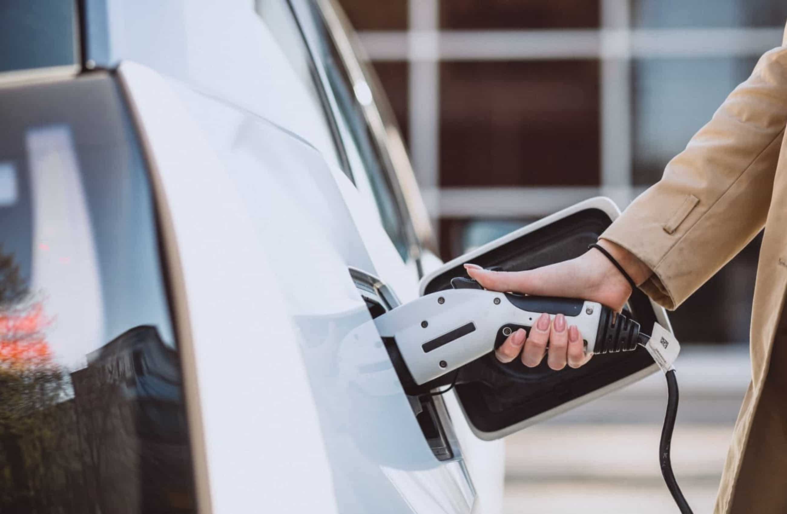 Mão de uma mulher segurando o carregador elétrico embutido em um carro branco.