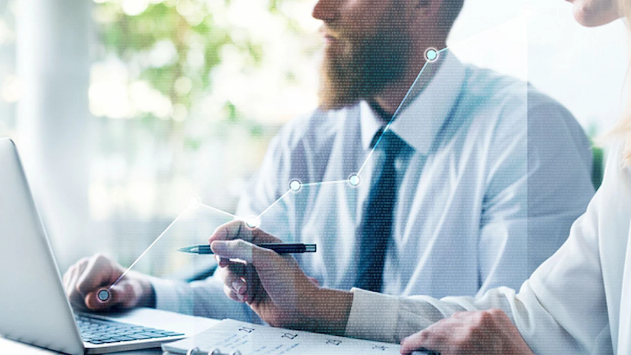 Um homem está sentado com o seu notebook. Ao seu lado, uma mulher com uma agenda na mesa e caneta na mão.