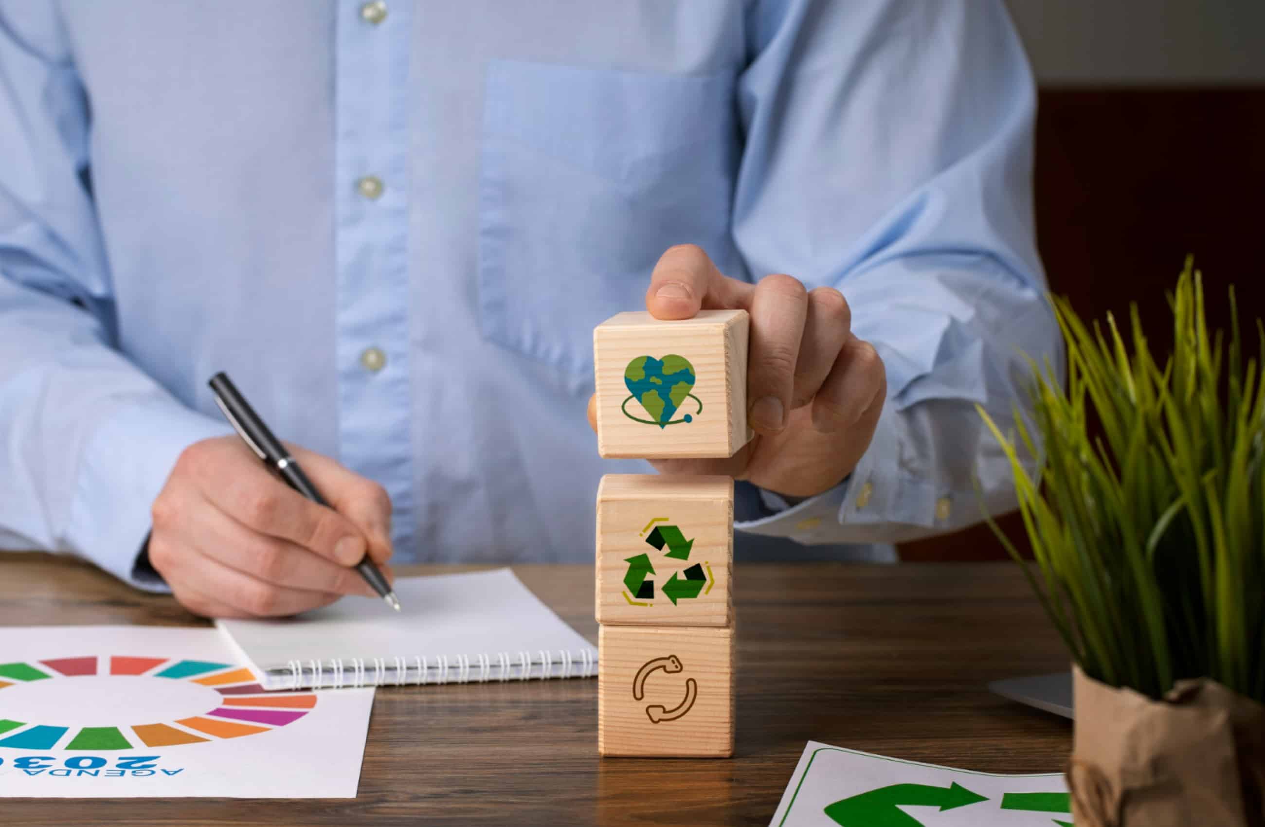 Pessoa empilhando blocos de madeira com símbolos de sustentabilidade e reciclagem em uma mesa de escritório.
