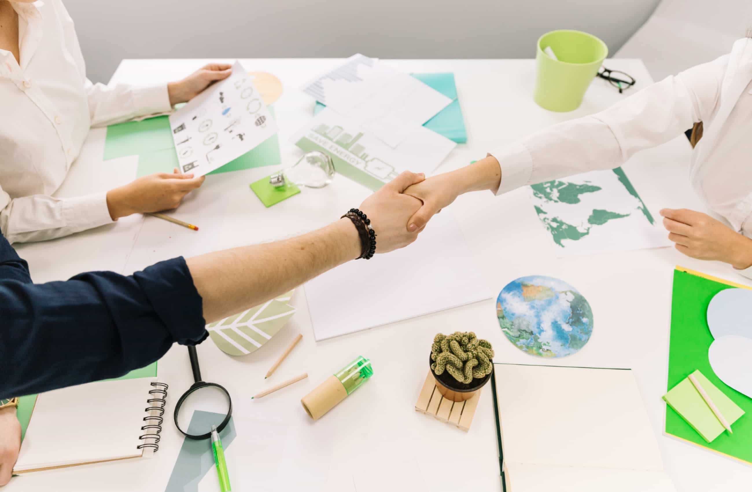 Duas pessoas apertando as mãos sobre uma mesa de trabalho com documentos, cadernos, e materiais de escritório focados em sustentabilidade.