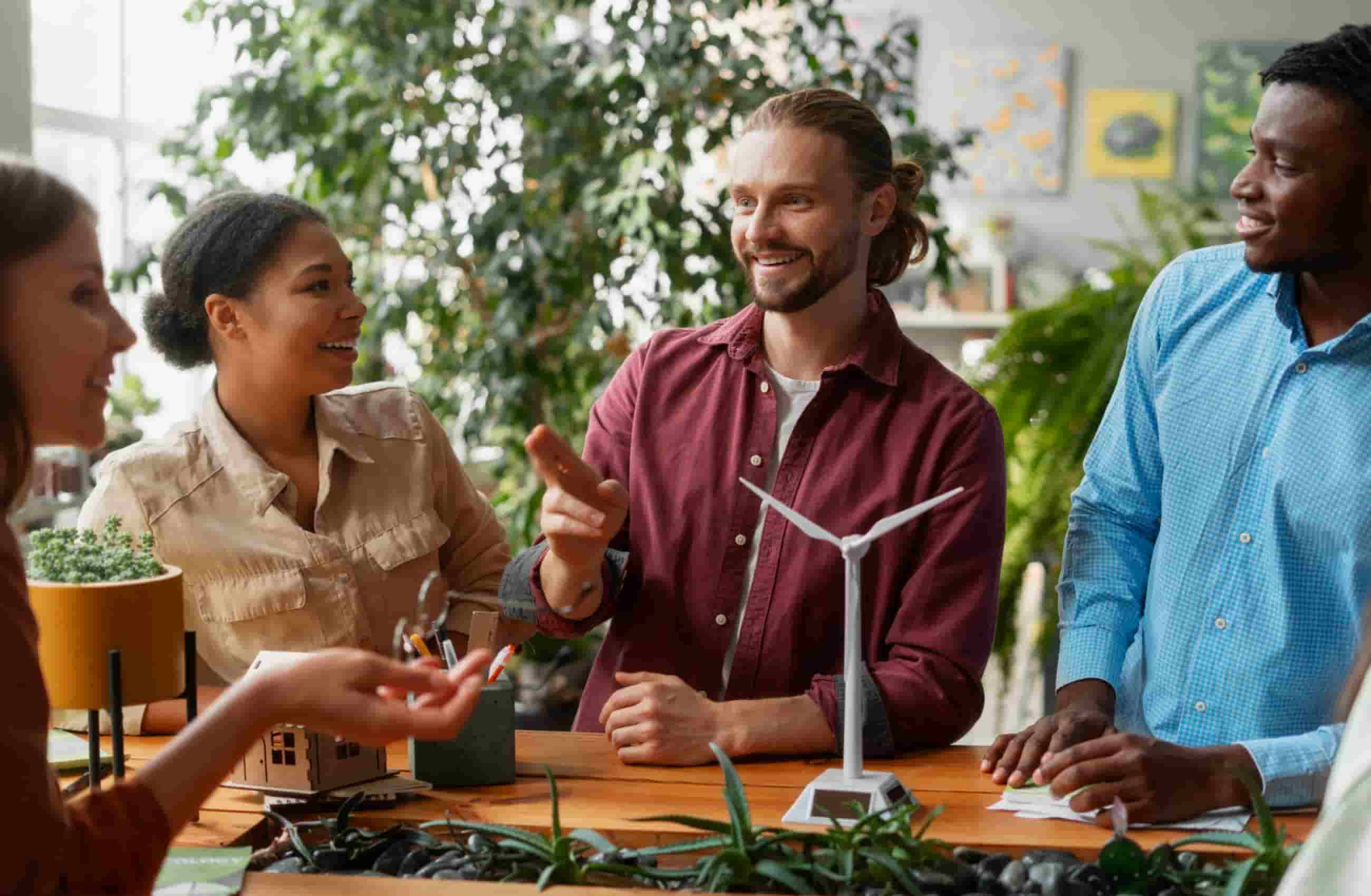 Grupo de pessoas discutindo ideias sobre energias renováveis, com um modelo de turbina eólica e plantas na mesa.