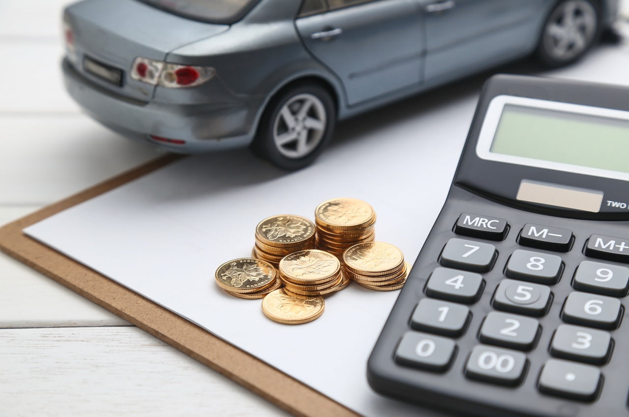 Miniatura de um carro sedan cinza disposto ao lado de moedas empilhadas e de uma calculadora.