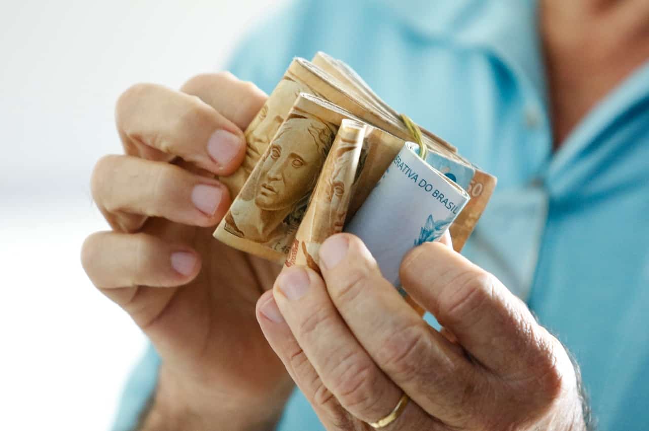 Mulher (seu rosto não aparece na imagem) segurando um bolo de dinheiro com notas de cem reais e cinquenta reais. Ela veste blusa azul básica.