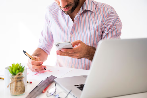 Um homem sentado em uma mesa de escritório enquanto usa um smartphone e escreve em uma folha de papel.