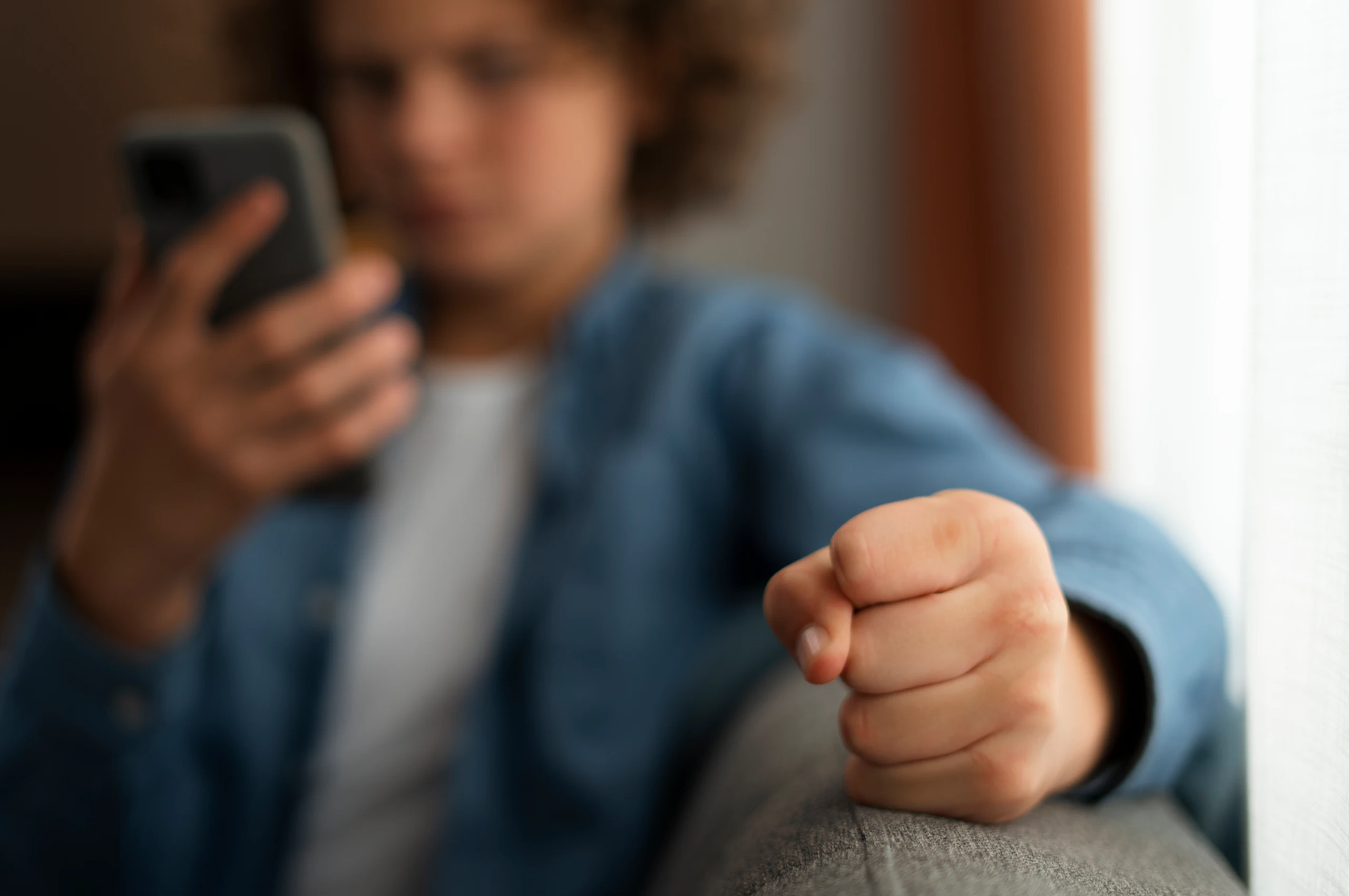Pessoa jovem segurando um smartphone na mão esquerda enquanto aponta o punho para a câmera, com o rosto desfocado ao fundo.