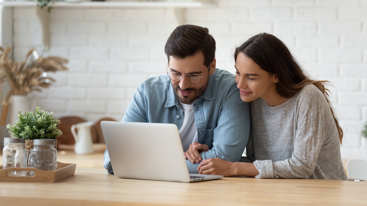 Casal sentado em frente a uma mesa, olhando para o notebook e conversando