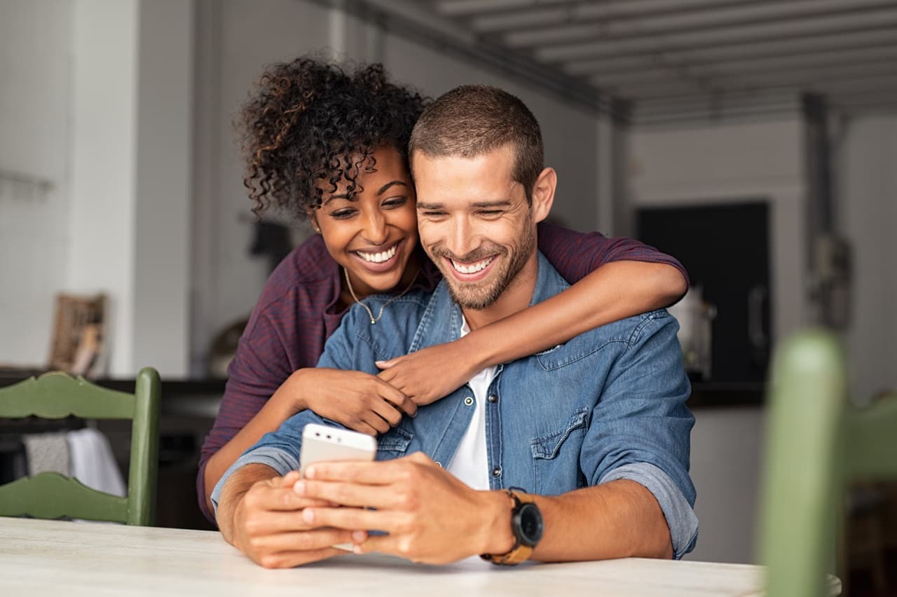 homem e mulher abraçados, sorrindo. O homem está segurando um celular nas mãos, os dois olham a tela do celular