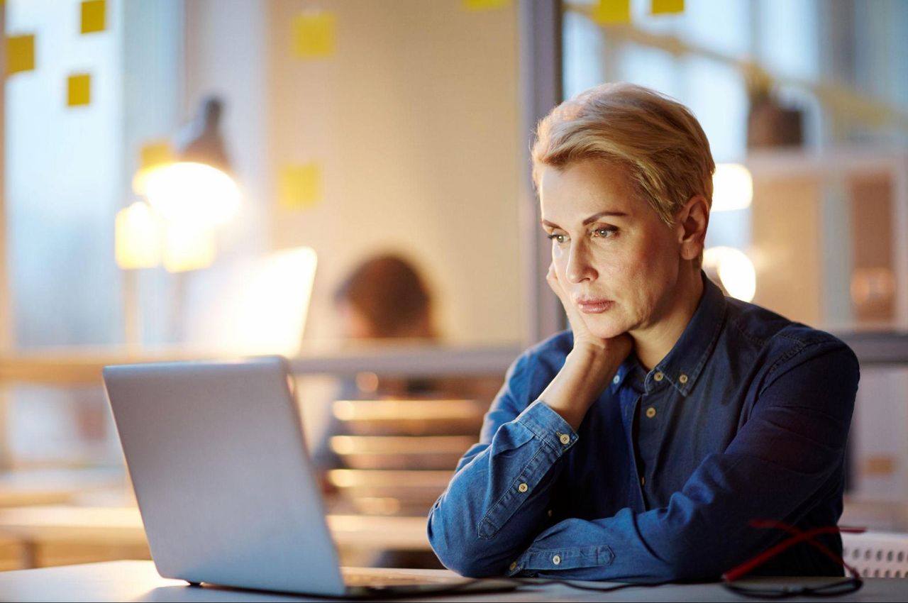 Mulher com expressão concentrada de cabelos curtos grisalhos veste blusa de botão jeans e faz entrevista de emprego online em laptop.