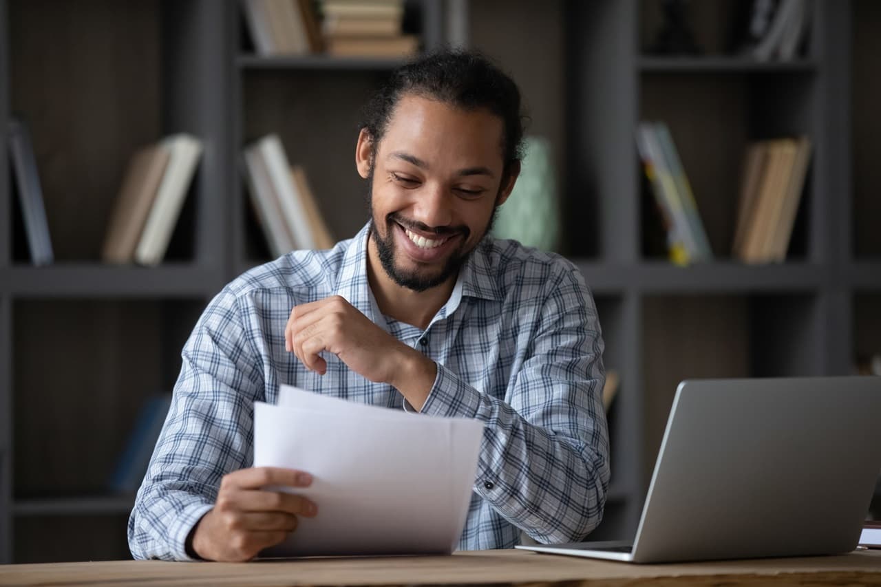 Pessoa sentada em uma mesa de trabalho analisando documentos impressos ao lado de um notebook aberto. Ao fundo, uma estante com livros compõe o ambiente interno.