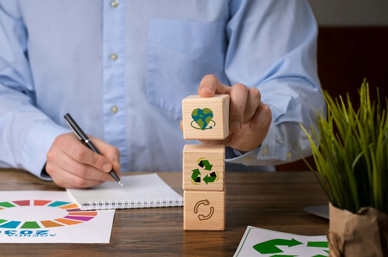 Pessoa empilhando blocos com símbolos de sustentabilidade em uma mesa, fazendo anotações.
