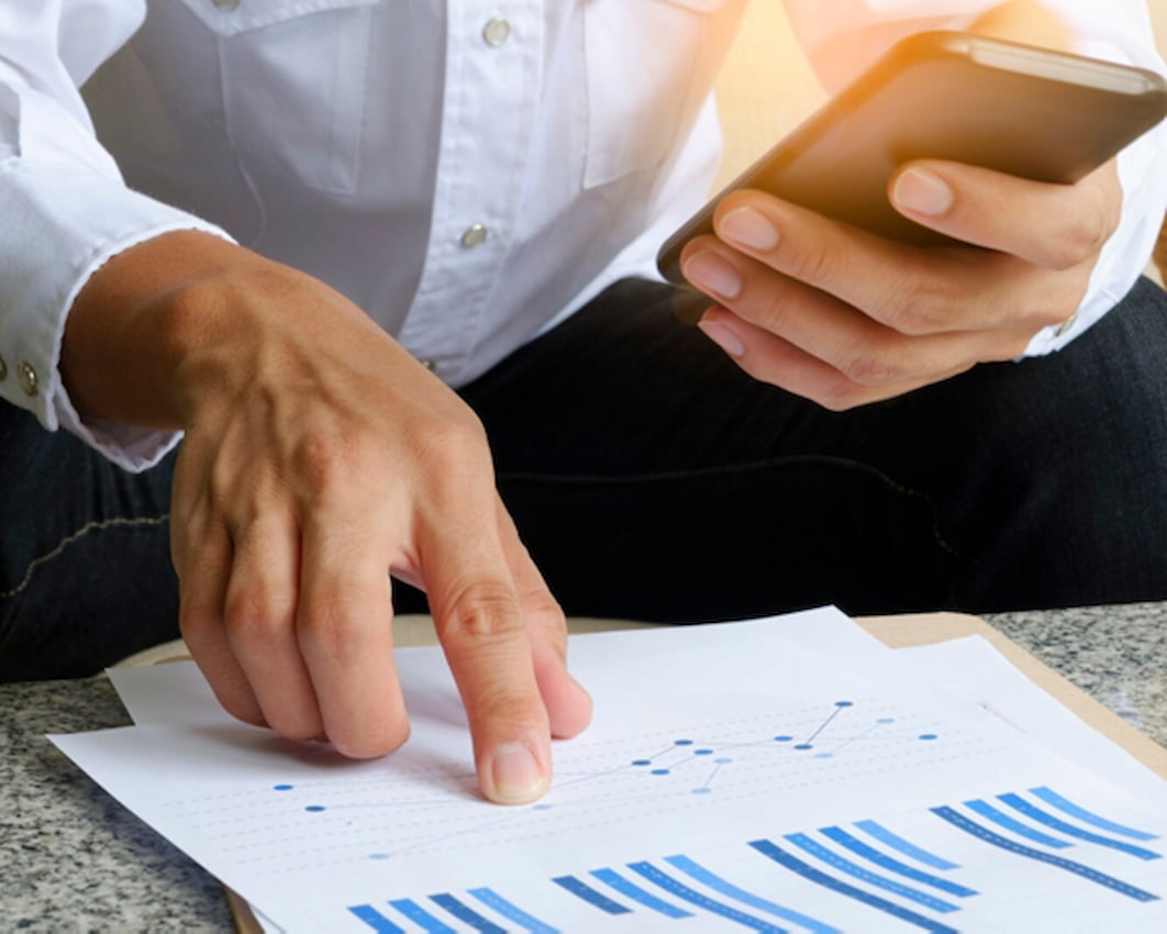 Pessoa analisando gráficos financeiros impressos enquanto consulta o celular, em contexto de planejamento e organização das finanças.